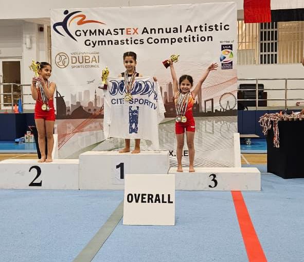 Our KG3 Young gymnast Julia Maroun holding her medals after achieving multiple top results at the Gymnastex Dubai gymnastics competition, standing inside a sports hall with gymnastics equipment in the background.