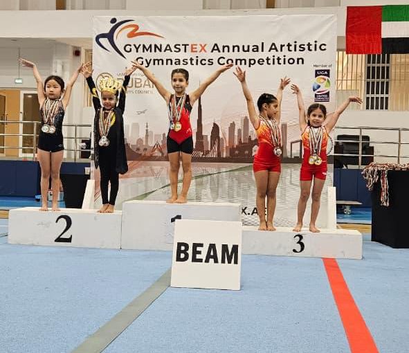 Our KG3 Young gymnast Julia Maroun holding her medals after achieving multiple top results at the Gymnastex Dubai gymnastics competition, standing inside a sports hall with gymnastics equipment in the background.