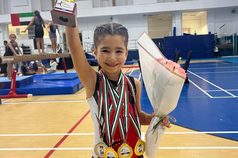 Our KG3 Young gymnast Julia Maroun holding her medals after achieving multiple top results at the Gymnastex Dubai gymnastics competition, standing inside a sports hall with gymnastics equipment in the background.