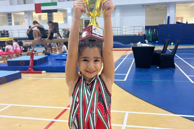 Our KG3 Young gymnast Julia Maroun holding her medals after achieving multiple top results at the Gymnastex Dubai gymnastics competition, standing inside a sports hall with gymnastics equipment in the background.