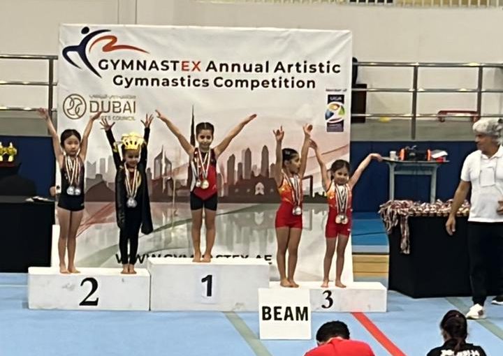 Our KG3 Young gymnast Julia Maroun holding her medals after achieving multiple top results at the Gymnastex Dubai gymnastics competition, standing inside a sports hall with gymnastics equipment in the background.